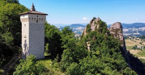 A tower with a cross on top, surrounded by lush greenery and mountains in the background. - Il Castellaro ai Sassi (Guiglia)