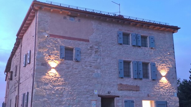 A stone building with multiple windows and doors, some with shutters, and a sign above the entrance. - Il Castellaro ai Sassi (Guiglia)