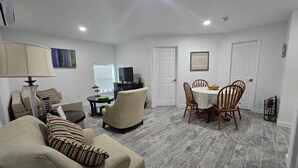 A living room with a sofa, armchair, and a dining table with chairs. There is a TV and a small table with a lamp.