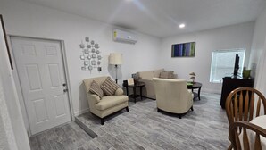 A living room with a sofa, armchair, and a small table with a lamp. There is a door, an air conditioning unit, and a window with blinds.