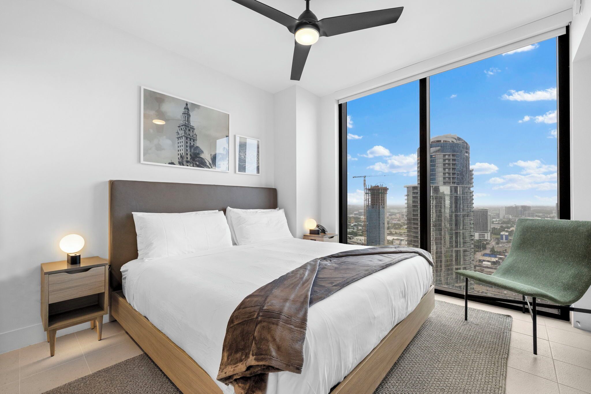 A modern bedroom with a large bed, a ceiling fan, a nightstand with a lamp, and a view of a cityscape through large windows.