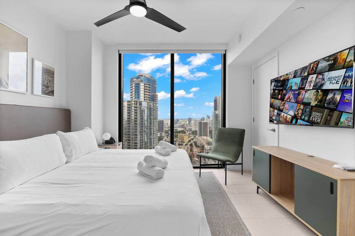 Ein modernes Hotelzimmer mit einem großen Bett, Blick auf die Stadt, Deckenventilator und einem an der Wand montierten Fernseher, der Filmplakate zeigt.