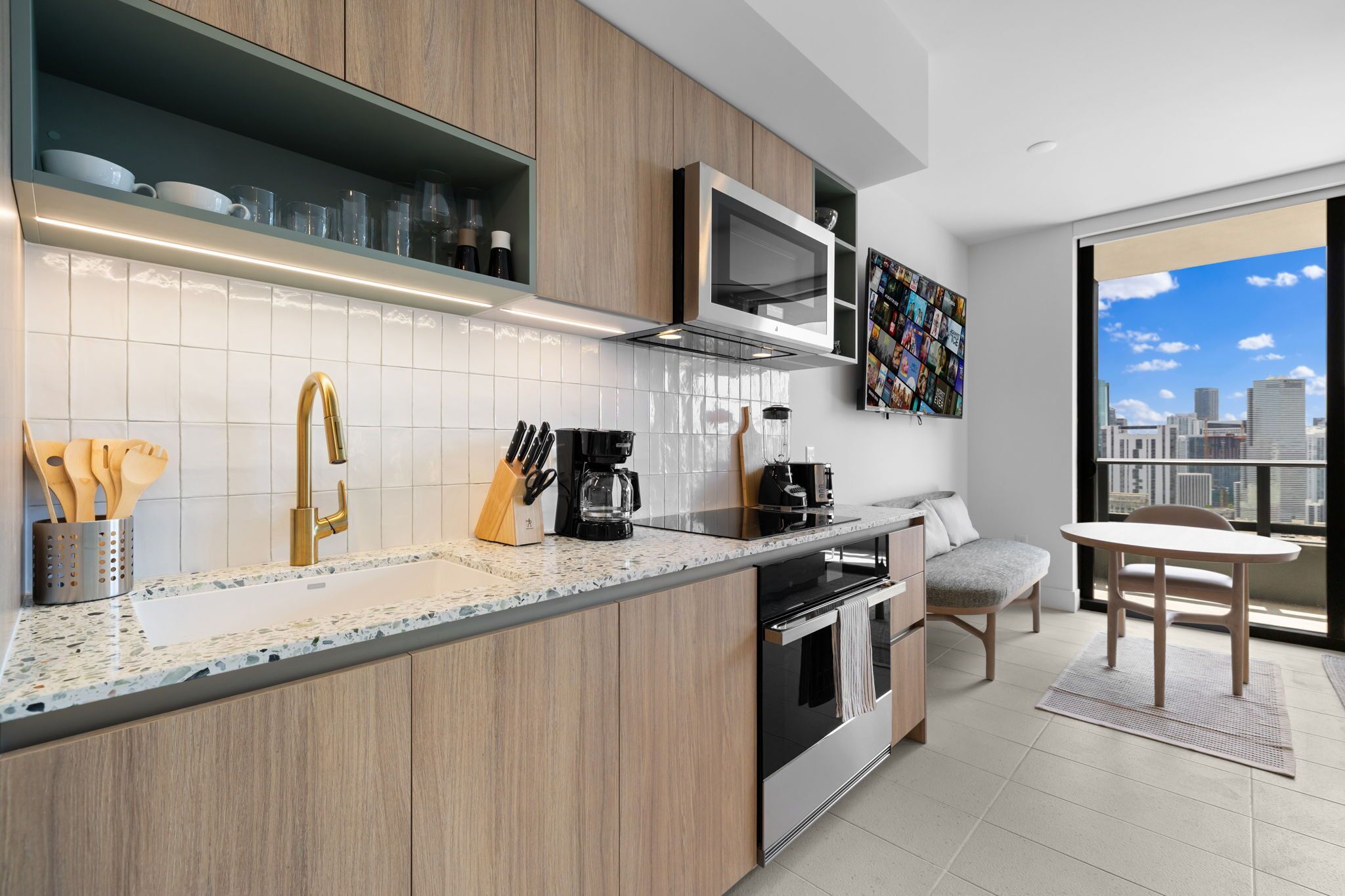 Una cocina moderna con armarios de madera, un lavamanos de acero inoxidable y un microondas integrado. Hay una zona de comedor con una mesa y sillas, y un ventanal con vistas a la ciudad.