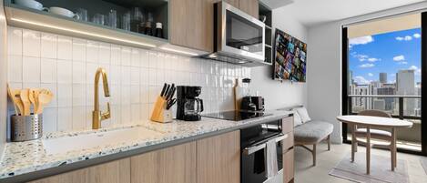 Una cocina moderna con armarios de madera, un lavamanos de acero inoxidable y un microondas integrado. Hay una zona de comedor con una mesa y sillas, y un ventanal con vistas a la ciudad.