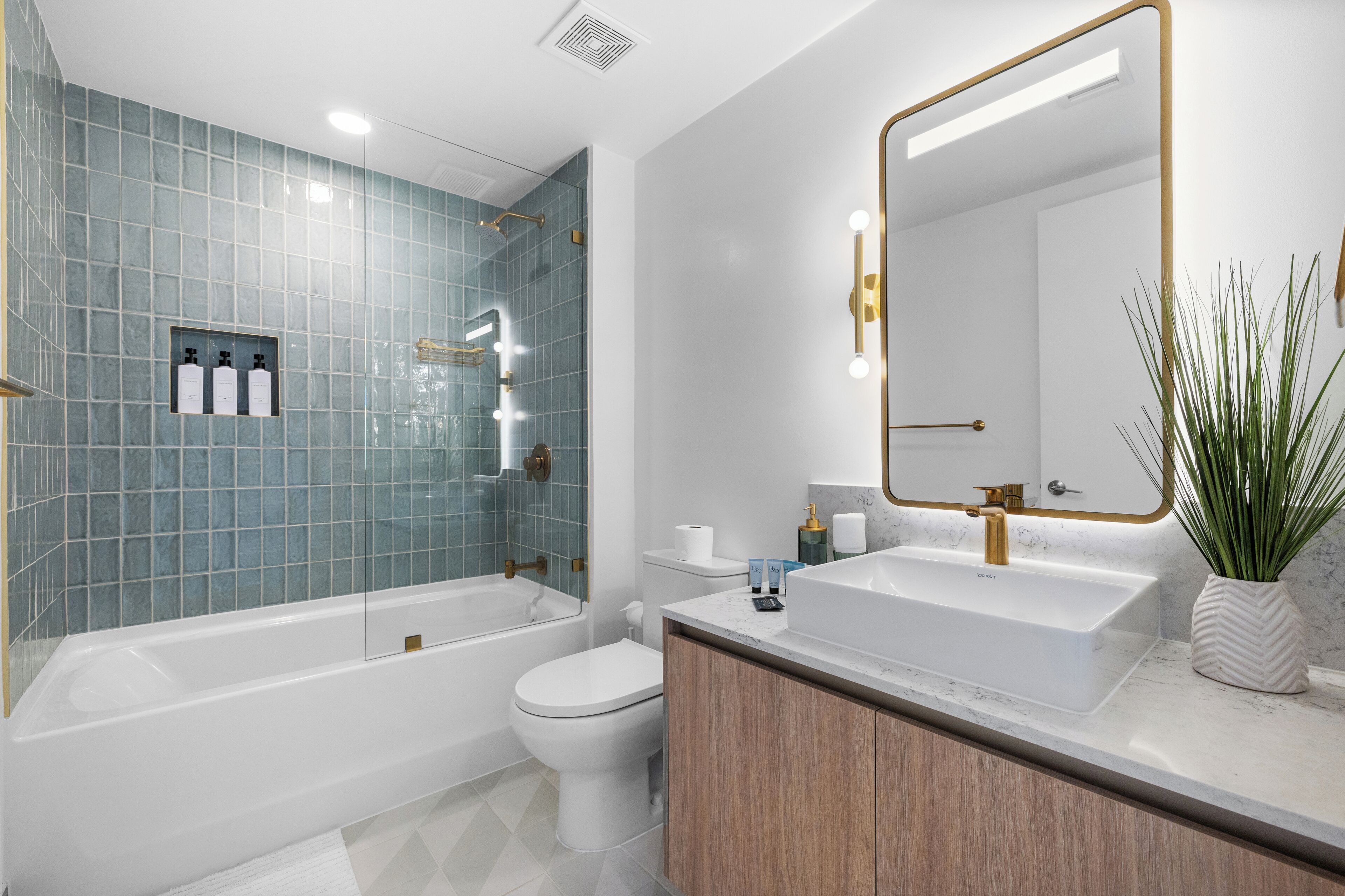 Ein modernes Badezimmer mit Badewanne, Toilette und Waschbecken, ausgestattet mit blauen Fliesen, einem großen Spiegel und einer Topfpflanze.