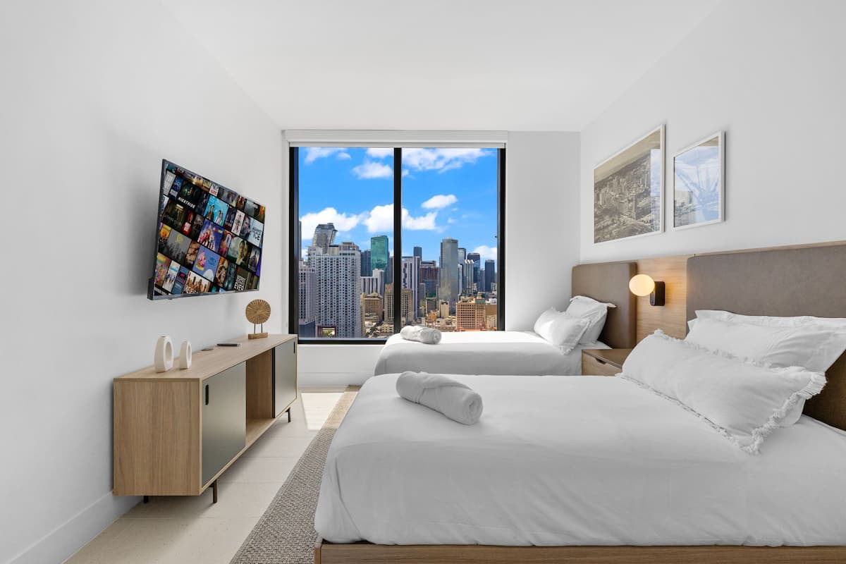 Ein modernes Hotelzimmer mit zwei Betten, einem großen Fernseher an der Wand und Blick auf die Stadt.