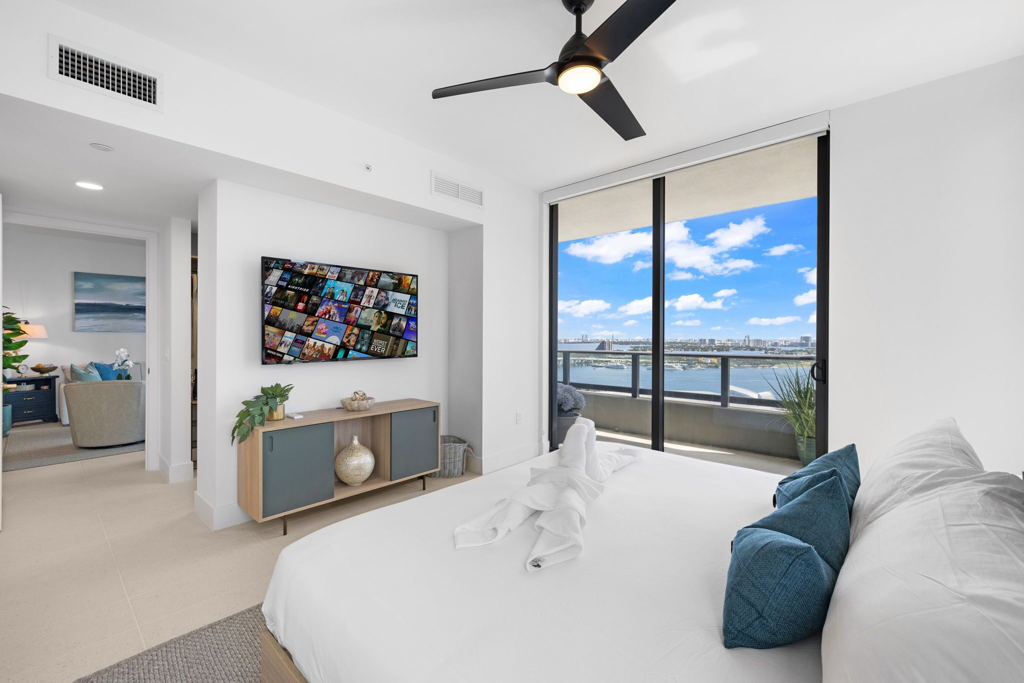 A modern bedroom with a large bed, a ceiling fan, a TV mounted on the wall, and a balcony with a city view.