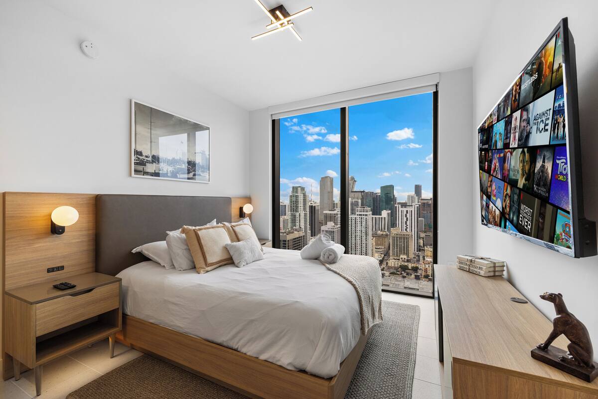 Ein modernes Schlafzimmer mit Blick auf die Stadt, ein großes Bett, Nachttische und ein wandmontierter Fernseher.