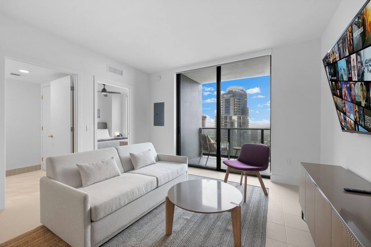 Ein modernes Wohnzimmer mit einer Couch, einem Couchtisch und einem Sessel. Ein großes Fenster bietet Blick auf eine Stadtlandschaft.