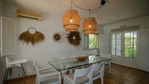 Un coin repas avec une table en verre, des chaises blanches et un bol. On y trouve deux lampes suspendues en osier, un climatiseur fixé au mur et une fenêtre avec des stores.