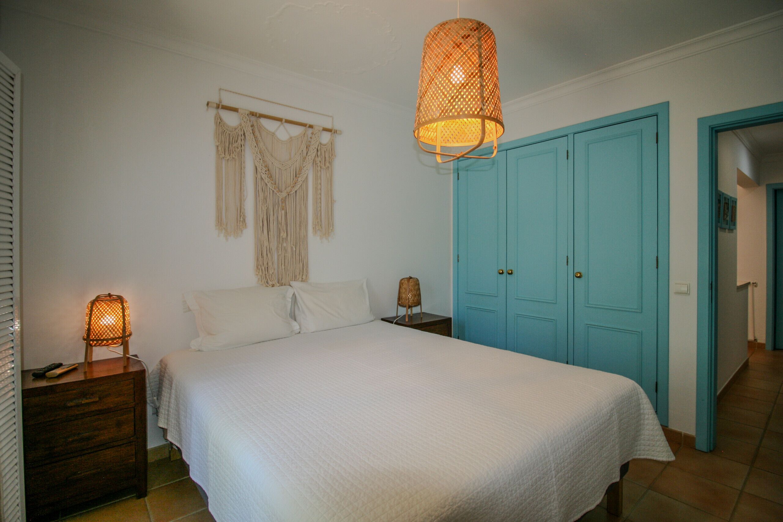 A bedroom with a bed, a nightstand, a lamp, macramé wall hanging, and a blue door.