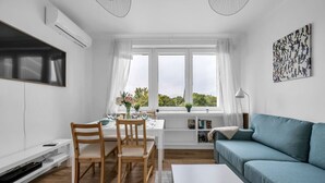 A modern living room with a blue sofa, a dining table set for four, and a large window with curtains.