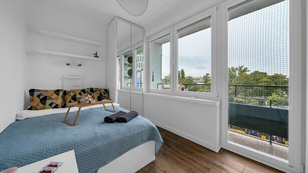A modern bedroom with a large bed, a side table, a mirror, and a view of the city through large windows.