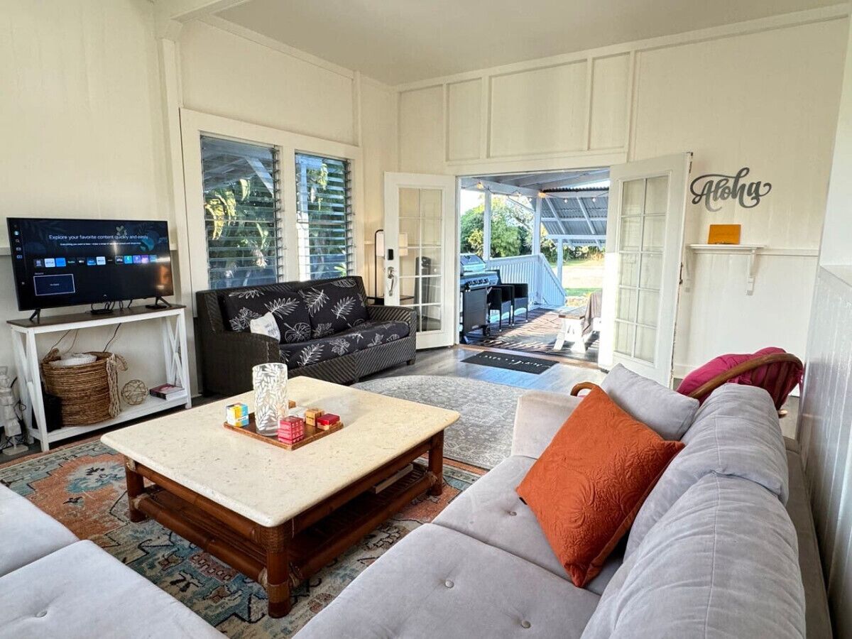 A spacious living room with a large sofa, a coffee table, and a television. There is a sliding glass door leading to an outdoor area.