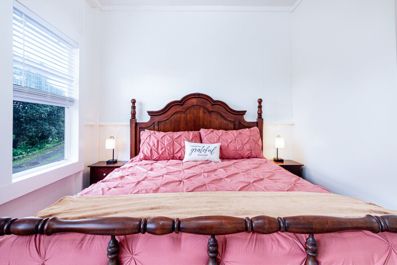 A neatly made bed with a wooden headboard, pink bedding, and a pillow with a message. There are two bedside lamps and a window with blinds.