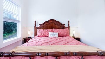 A neatly made bed with a wooden headboard, pink bedding, and a pillow with a message. There are two bedside lamps and a window with blinds.