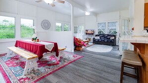 A spacious living area with a large window, a ceiling fan, and a dining area with a table and bench. - Your Perfect Hilo Homebase - Sleeps 9 with AC (Hilo)