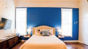 Quarto com cama grande, duas mesas de cabeceira, TV na parede e janela com persianas.