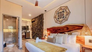 A modern bedroom with a large bed, a wooden headboard, a wall-mounted artwork, and a bathroom visible through a glass door. - Amira Itzamna Luxe Studio Close to beach! (Tulum)
