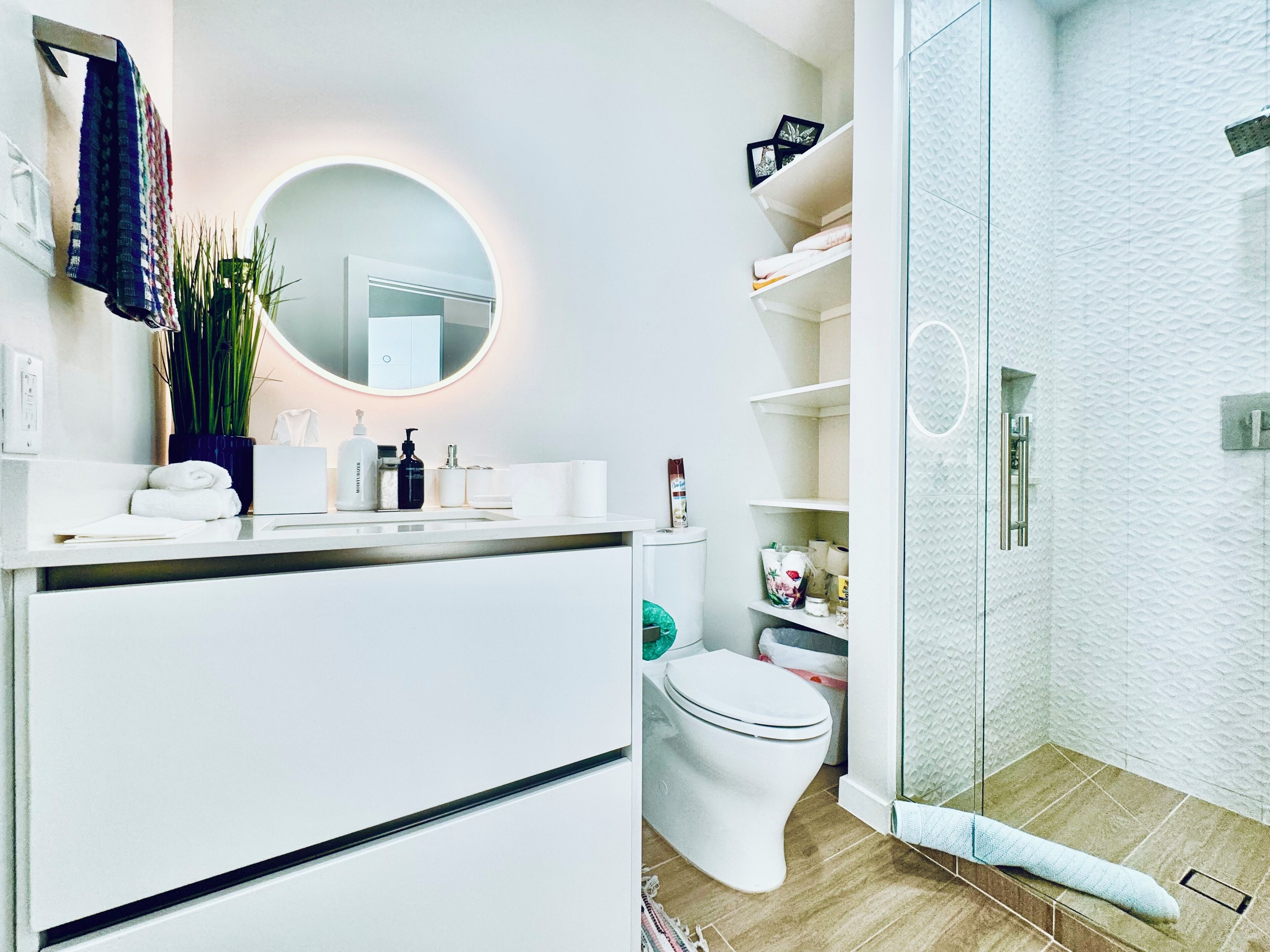 A modern bathroom with a white vanity, a round mirror, a toilet, and a glass-enclosed shower area.