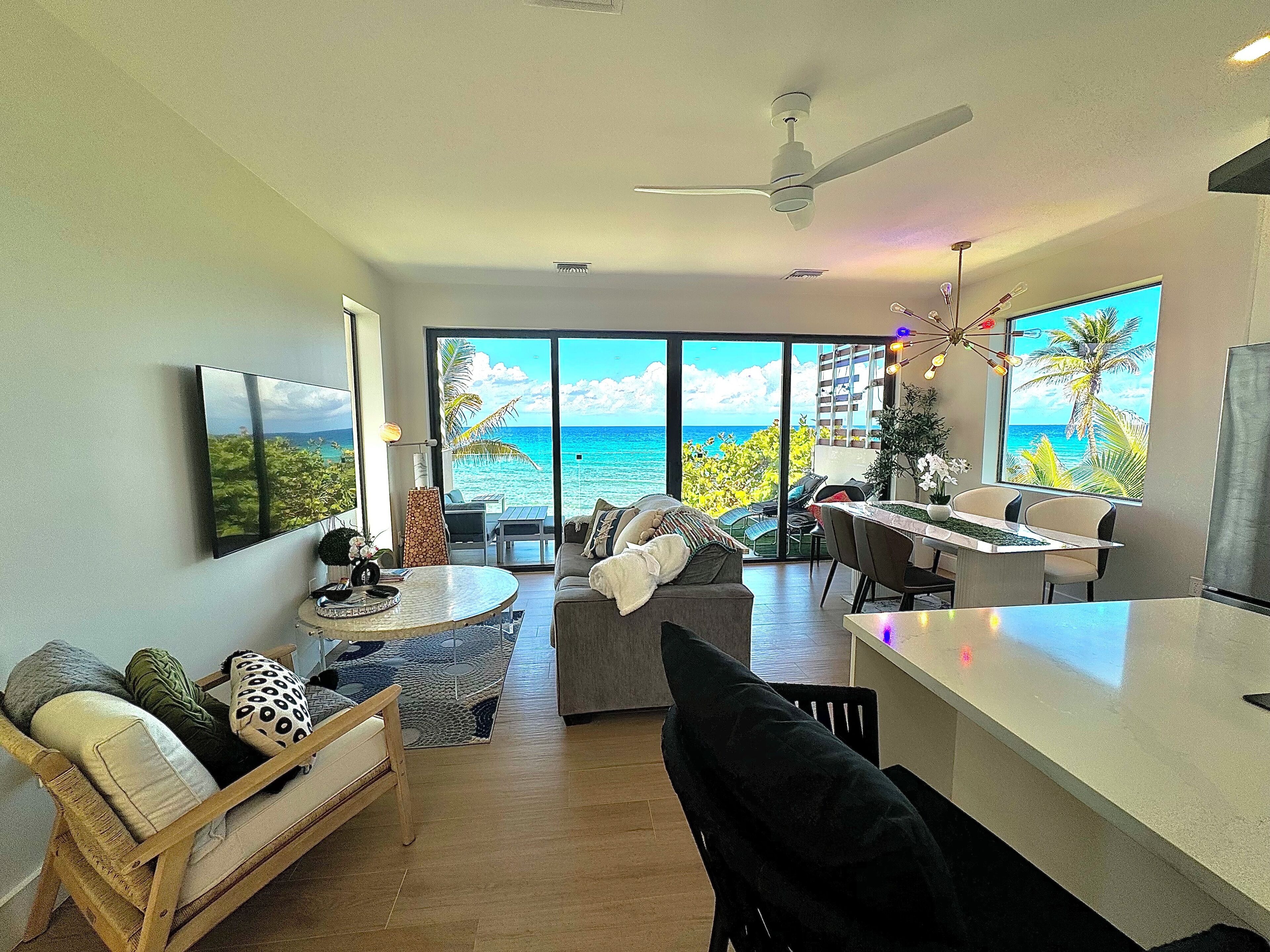 A modern living room with a large window overlooking the ocean, a ceiling fan, a dining area, and a kitchen island.