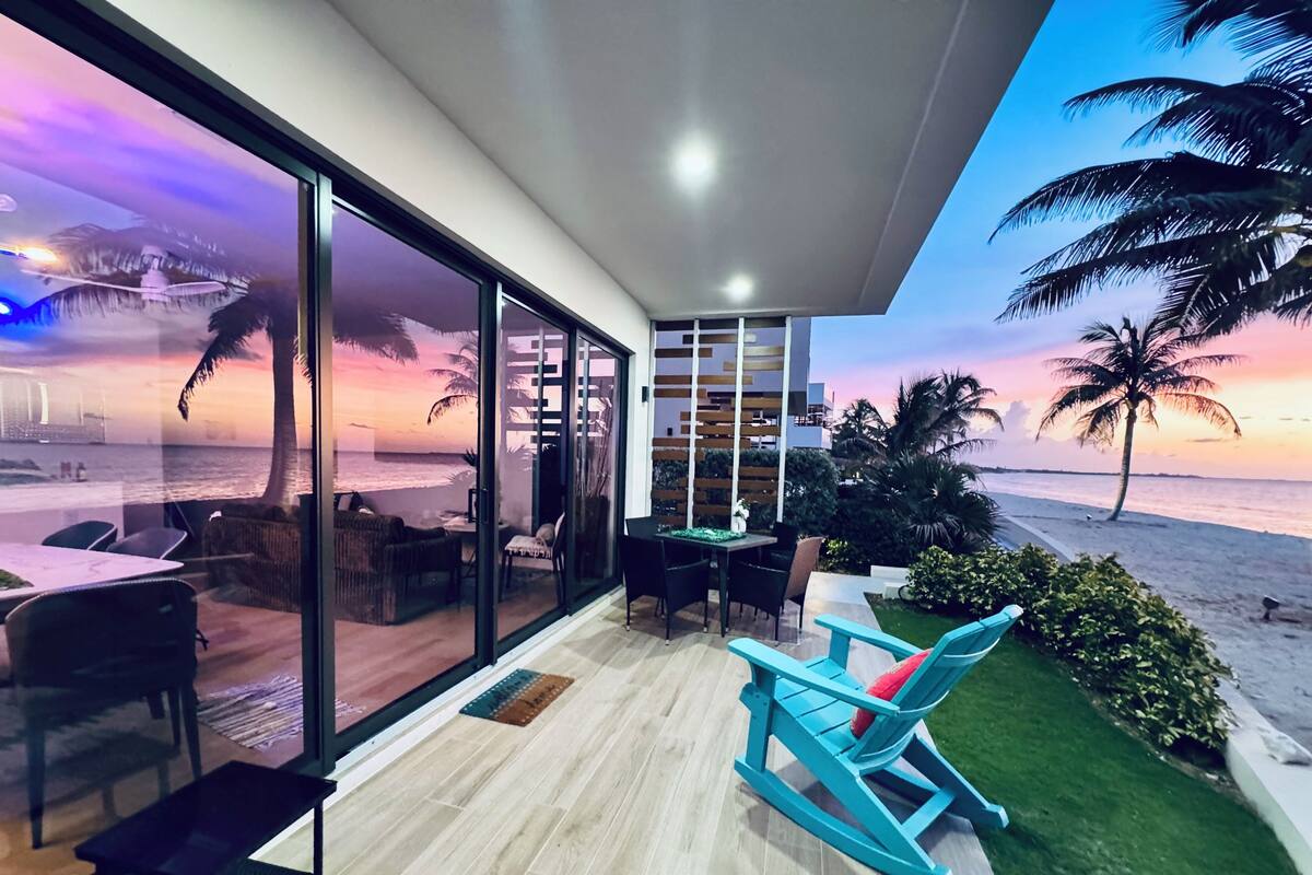 A modern room with a large sliding glass door leading to a beach view. Inside, there is a dining area with a table and chairs, a small seating area with a rocking chair, and a small table with a vase.