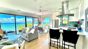 A modern living room with a large window overlooking the ocean, a dining area with a kitchen in the background, and a ceiling fan. - Silver Reef Residencies  (North Side)