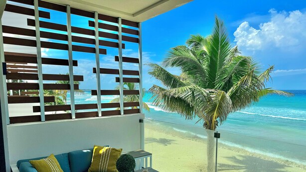 A balcony with a view of the beach, a sofa with cushions, a small table with a plant and seashell, and a palm tree. - Silver Reef Residencies  (North Side)