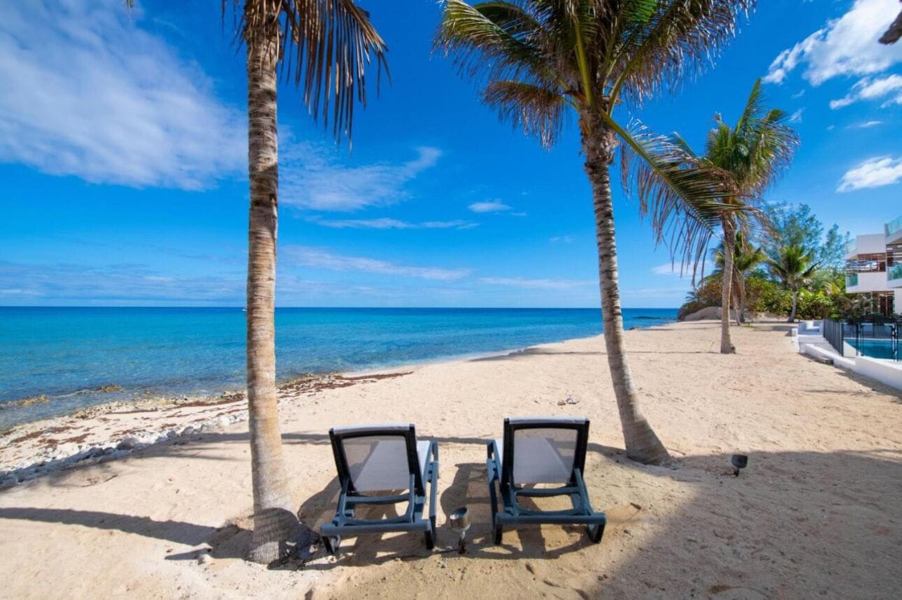 A beach with two lounge chairs, palm trees, and a clear blue sky.