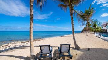 A beach with two lounge chairs, palm trees, and a clear blue sky.