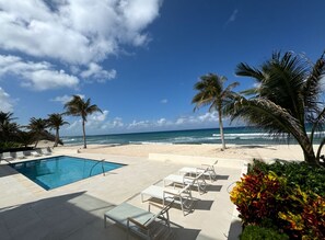 Ein Poolbereich mit Liegestühlen und Palmen, mit Blick auf einen Strand und das Meer.