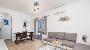 Ett modernt vardagsrum med en soffa, ett matbord och stolar. Det finns ett litet runt bord med en växt på. Rummet har ett stort fönster med gardiner, en luftkonditioneringsanläggning och en väggmonterad lampa.