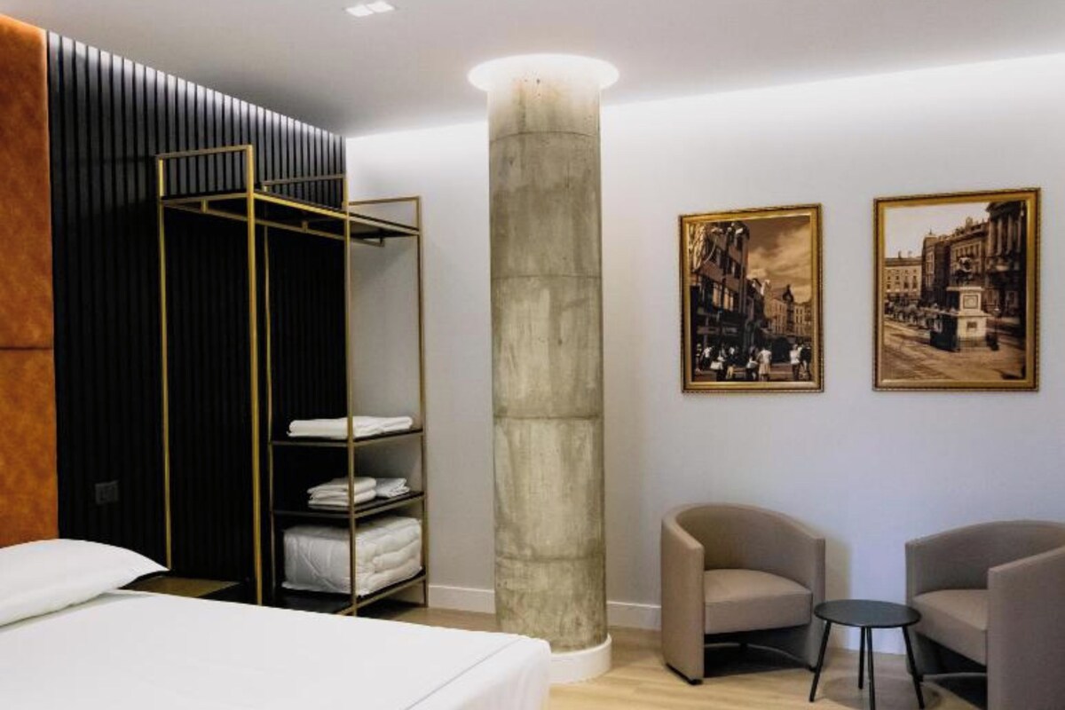 Ein modernes Hotelzimmer mit einem Bett, einem Kleiderschrank mit darin hängender Kleidung, einer Sitzecke mit zwei Sesseln und zwei gerahmten Bildern an der Wand.