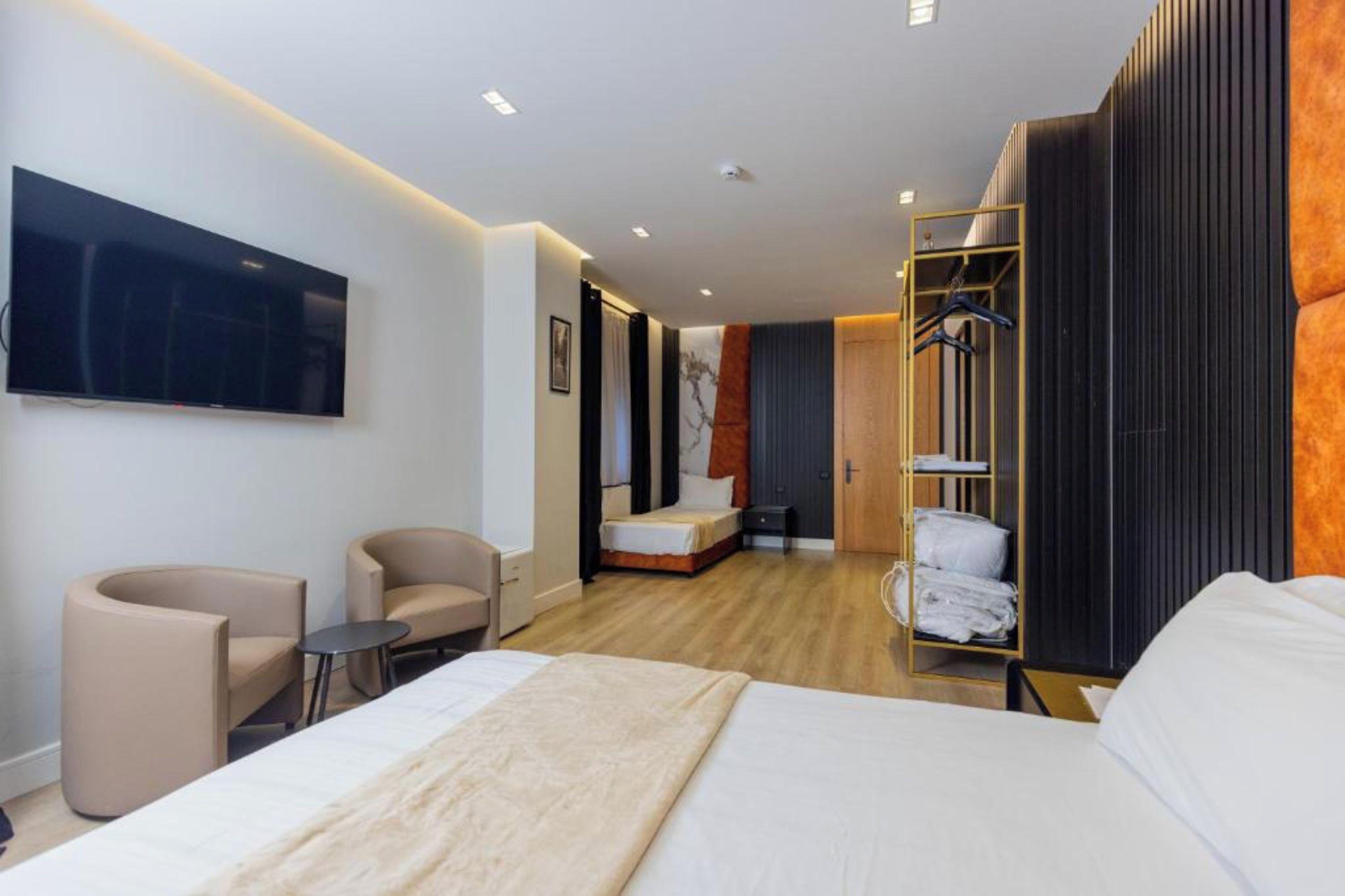 A modern hotel room with a large bed, two armchairs, a small table, a TV mounted on the wall, and a walk-in closet with a mirror.