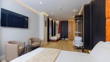 A modern hotel room with a large bed, two armchairs, a small table, a TV mounted on the wall, and a walk-in closet with a mirror.