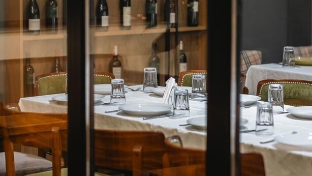 Een restaurant met meerdere tafels klaar voor diners, een wijnrek met diverse flessen en uitzicht door een glazen scheiding.