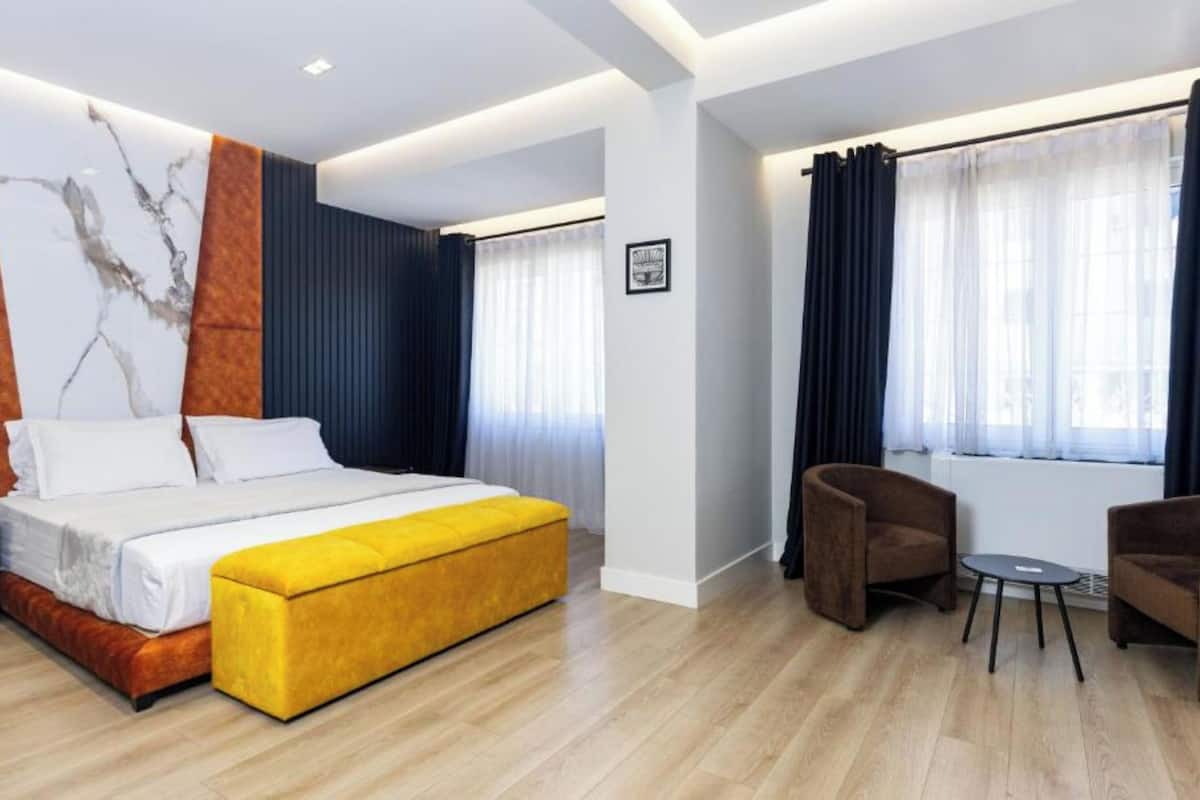Una habitación de hotel moderna con una cama grande, un reposapiés amarillo, un sillón marrón y una mesita pequeña.