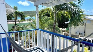 A balcony with a hammock, white railings, and a view of a green building and palm trees. - Beautiful ocean view 4-bedroom house with AC in the island of Culebra PR (Culebra)
