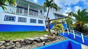 A two-story building with a blue and white exterior, a balcony with white railings, and a stone pathway leading to the entrance. - Beautiful ocean view 4-bedroom house with AC in the island of Culebra PR (Culebra)