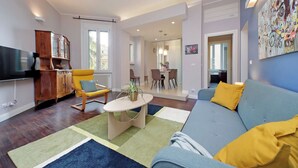 A modern living room with a sofa, armchair, wooden cabinet, and a television. - 4BNB - Armot House Vatican Apartment (Rome)