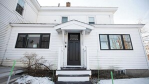 Une maison blanche à deux étages, avec une porte d’entrée noire, un petit porche et une cheminée.