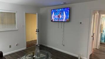 Una sala de estar con una televisión de pantalla plana montada en la pared, una mesa de centro de vidrio con objetos decorativos, un sofá marrón oscuro y piso de madera.