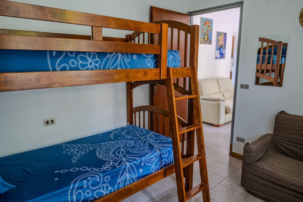 Ein Stockbett mit blauen Bettbezügen, ein Holzstuhl und ein Sofa in einem Zimmer mit Fliesenboden und Bildern an den Wänden.