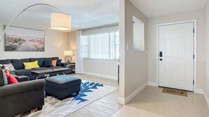 A modern living room with a sofa, ottoman, coffee table, and a large floor lamp. There is a door with a doormat and a window with blinds.