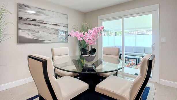 A dining area with a glass table surrounded by four chairs, a vase of pink flowers, and a beach scene painting on the wall.