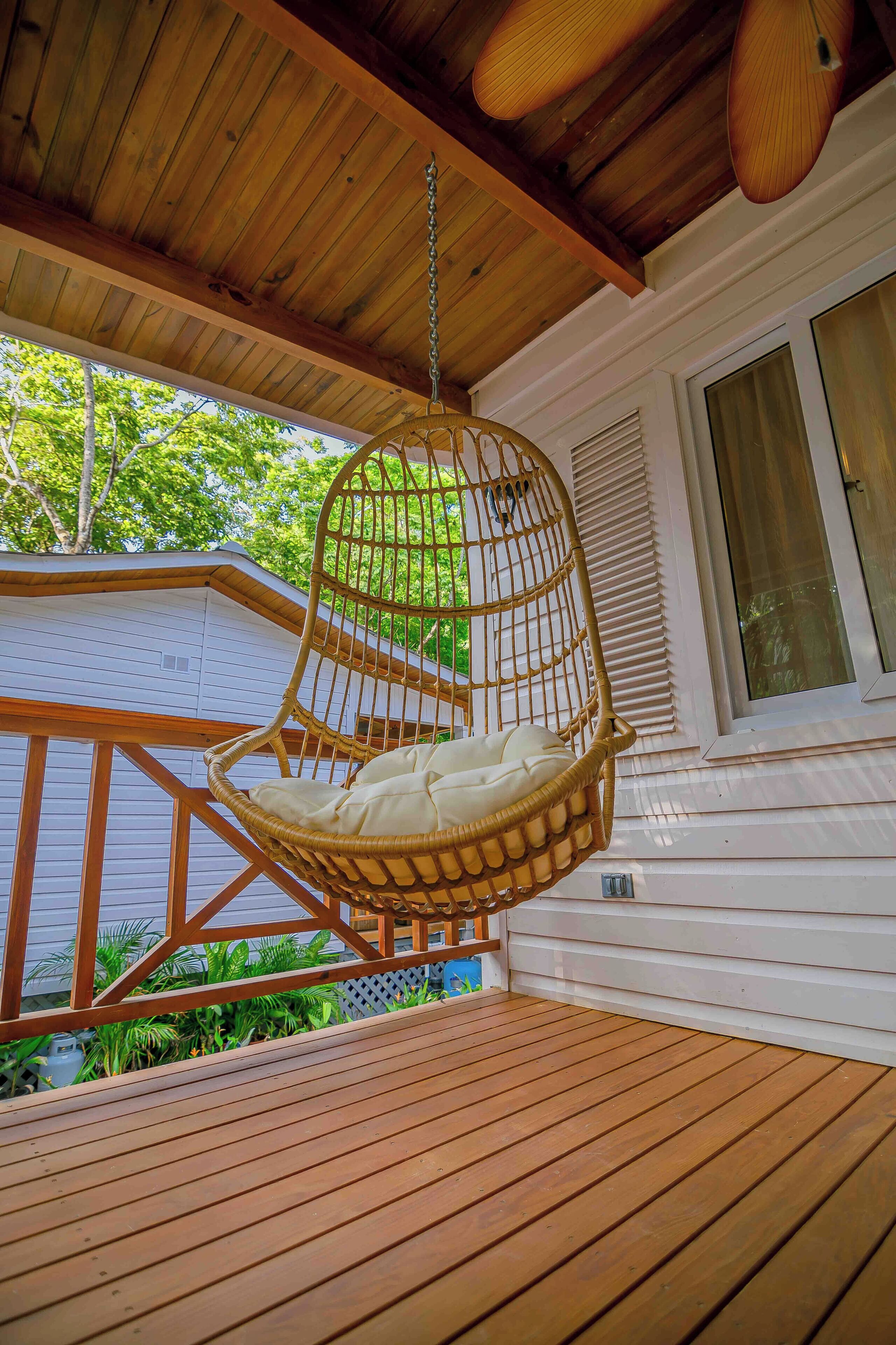 A wooden deck with a hanging wicker chair and a window with blinds.
