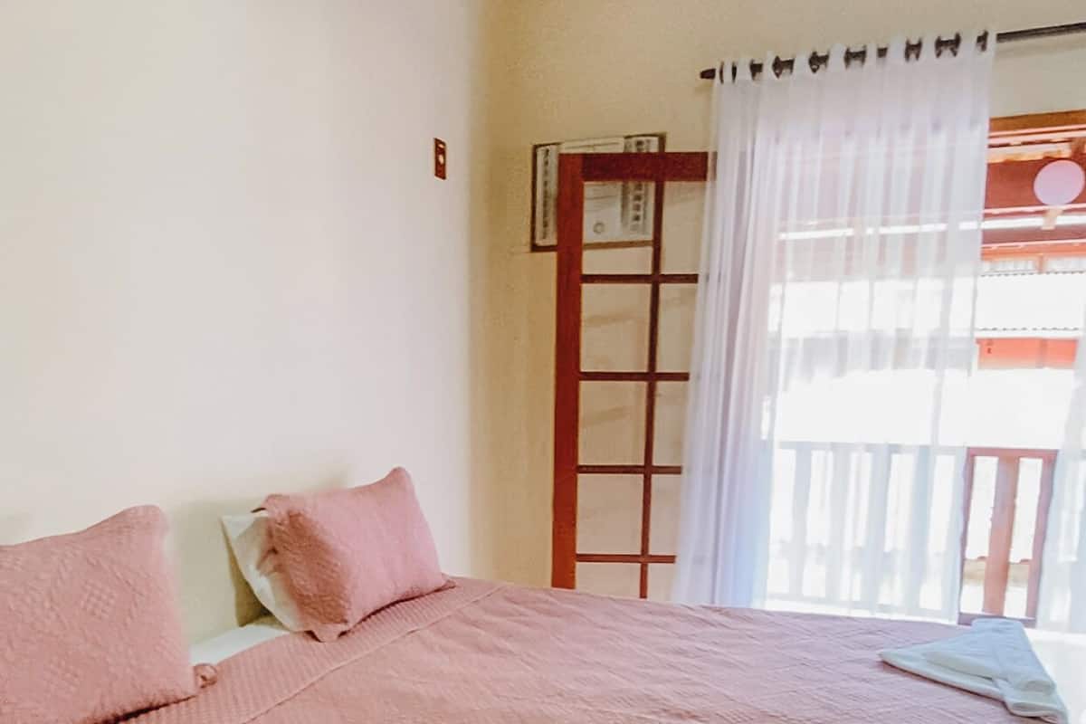A single-bed room with a wooden headboard, a window with a white curtain, and a view of a balcony.