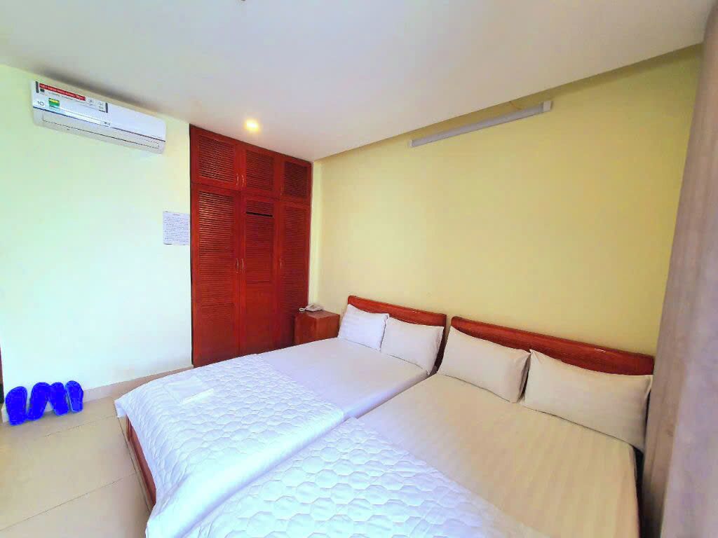 A hotel room with two beds, a wooden wardrobe, an air conditioning unit, and a pair of slippers.