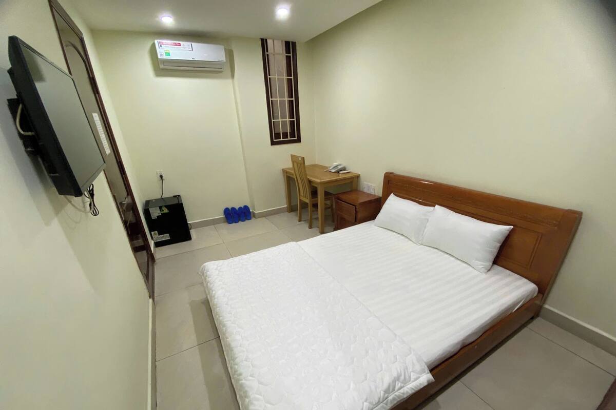 A hotel room with a large bed, a desk, a chair, a TV, and an air conditioning unit.