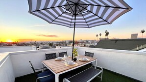 A rooftop terrace with a wooden table set for two, a striped umbrella, and a view of the city skyline at sunset.